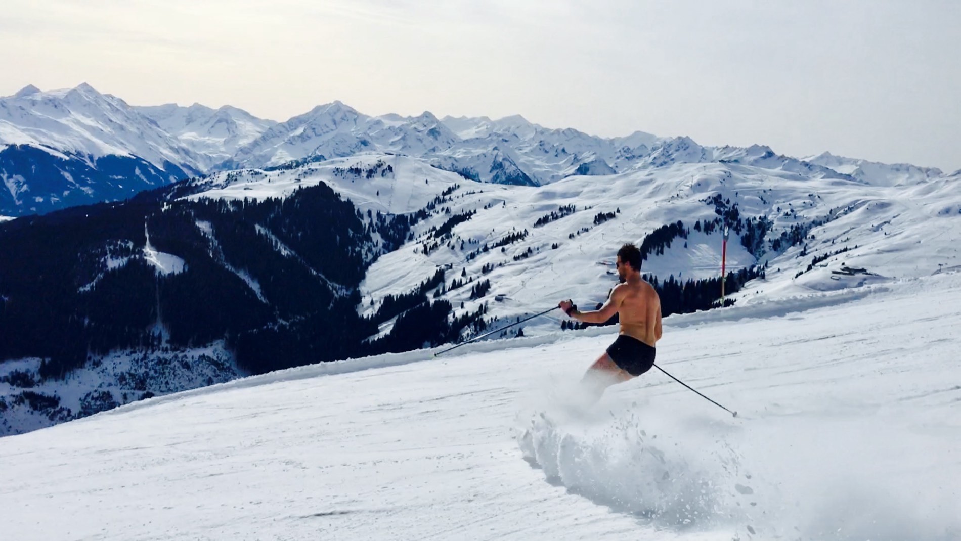 Max Gotzler skiing in swim shorts, snowy mountains behind him